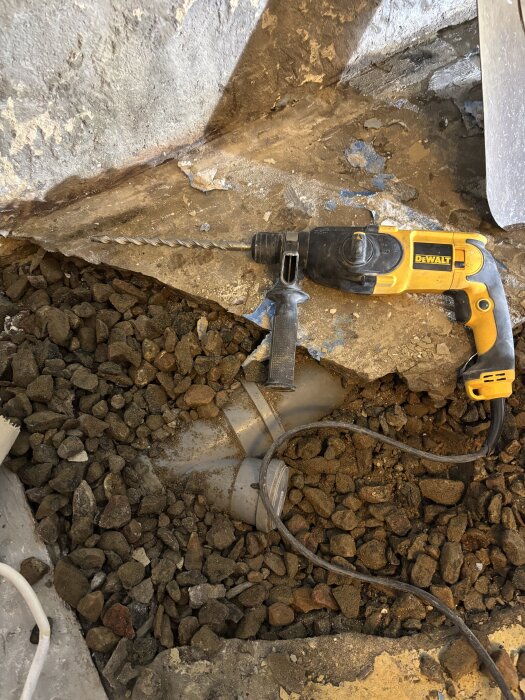 En gul borrmaskin på en byggarbetsplats med sten och gammalt duschbrunnslock synligt i marken.