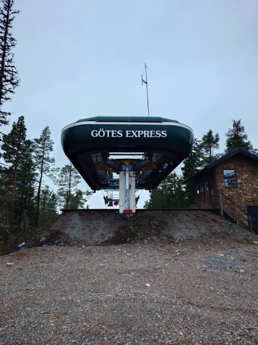 Skidlift märkt "GÖTES EXPRESS", med skog i bakgrunden och stuga till höger. Grå himmel och vinterlandskap.