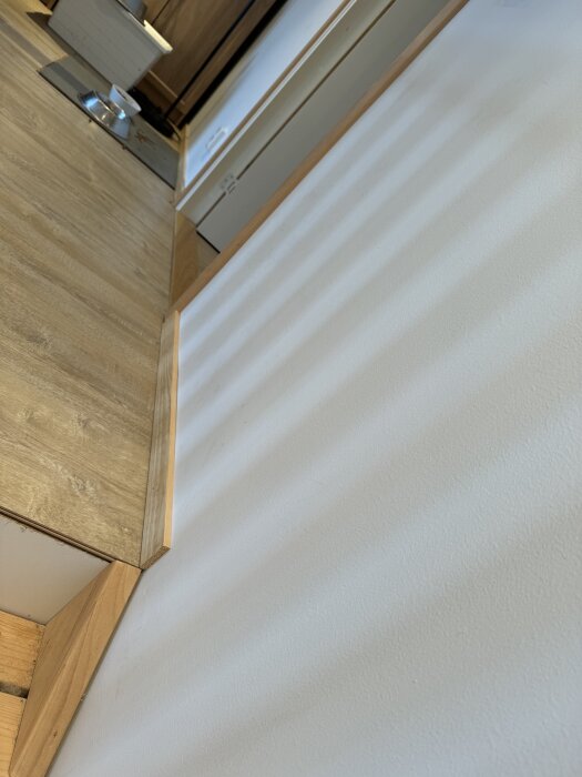 Vinklad bild av trä- och laminatgolv med nyligen monterade lister längs väggen, del av ett renoveringsprojekt hemma.