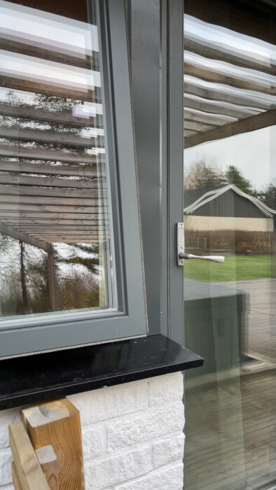 Grått fönster och dörr med Osby-glas, sedd utifrån med reflektioner av trädgård och pergola i glaset.