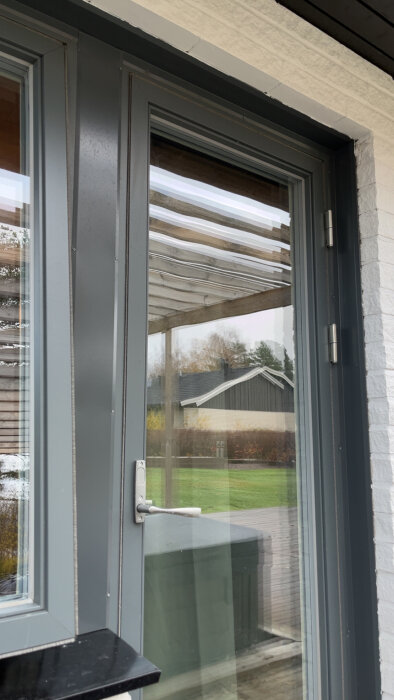 En grå glasdörr med metallhandtag och fönster i en vit fasad, utsikt över en trädgård och ett hus i bakgrunden.