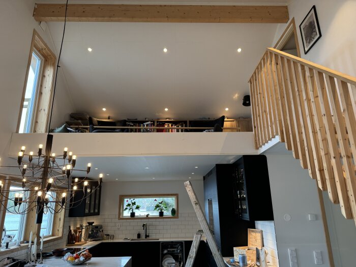 Modernt kök med vita väggar, trädetaljer och en stor ljuskrona. Ett loft med räcke och soffor syns ovanför. Stege och målargrejer på golv.