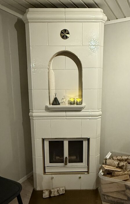 Vit kakelugn med två nedre reglage, ved i en behållare bredvid; dekorativ nisch med ljus ovanför.