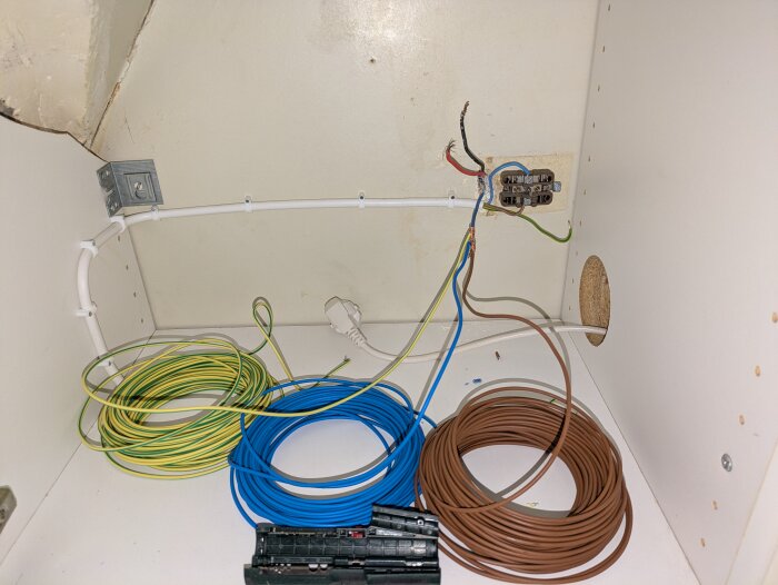 Gammal elinstallation med färgglada kablar i en garderob med vit inredning; röda, svarta och blåa trådar syns.