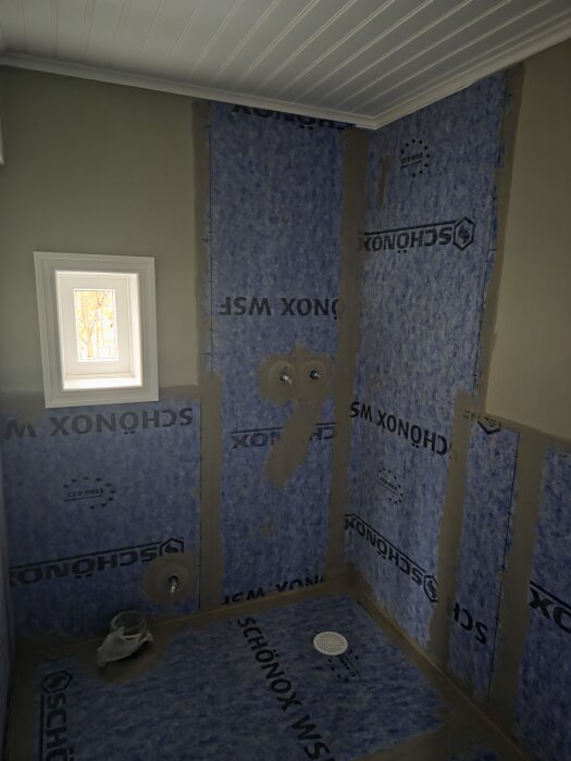 Badrumshörn klätt med blå SCHÖNOX vattentätningsfolie och monterad golvbrunn. Ett litet fönster sitter i väggen till vänster.