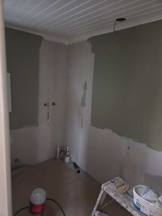 Ett rum under renovering med spacklade väggar och målarfärg, byggmaterial på golvet och en stege.