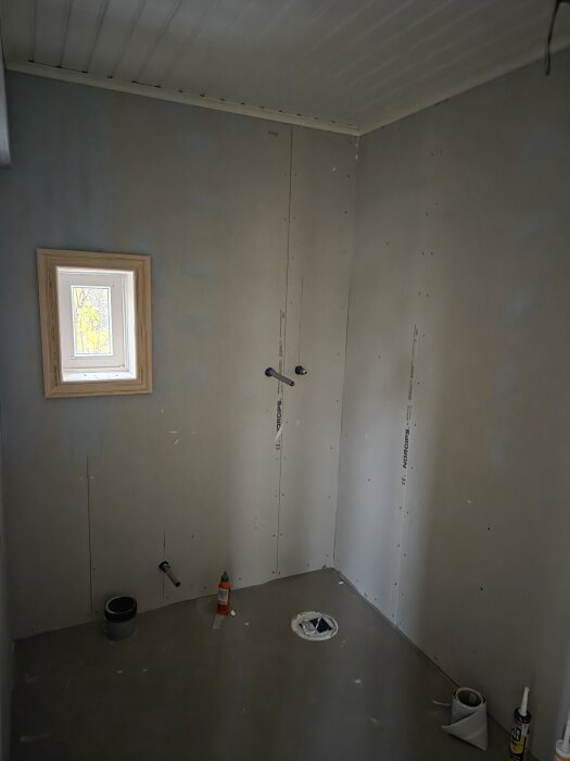 Rum under renovering med gipsskivor och målarfläckar på väggarna, litet fönster och rör för VVS-installation.