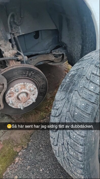 Närbild på bilens bromsskiva och borttaget hjul med dubbdäck, står på ojämn mark.