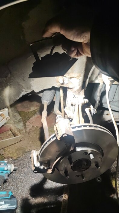 Bromsskiva och bromsok under reparation på bil, med fokus på vänster framhjul. Skugga av hand syns hålla verktyg ovanför delen.