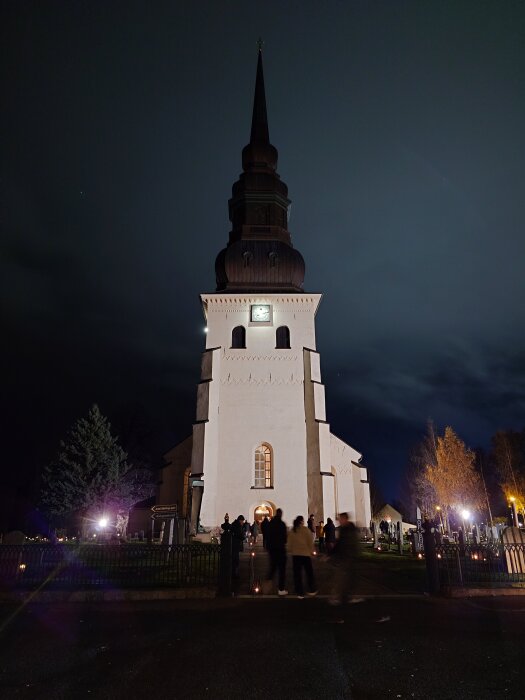 En kyrka med en hög tornspira upplyst på kvällen, omgiven av människor och träd.