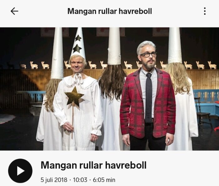 En grupp människor i traditionella luciakläder står på en scen. En av dem bär en rutig kavaj. Bakgrunden har dekorationer av träbockar.