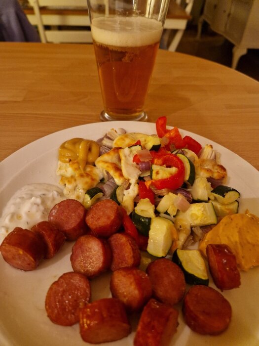 Tallrik med ugnsrostade grönsaker, korv och ost, tillsammans med tsatsiki och annan sås. Ett glas öl står på bordet.