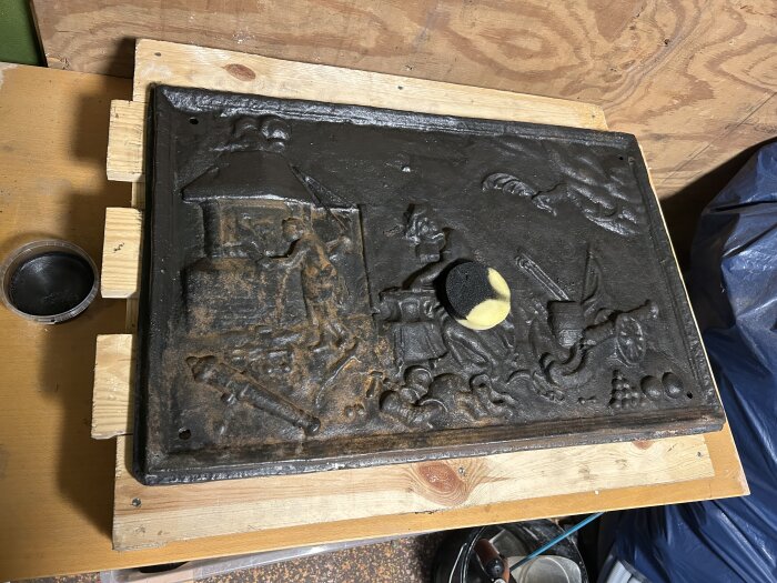 Järnspisplatta med reliefer av människor och symboler, nyligen behandlad med svart färg, en svamp ligger ovanpå.