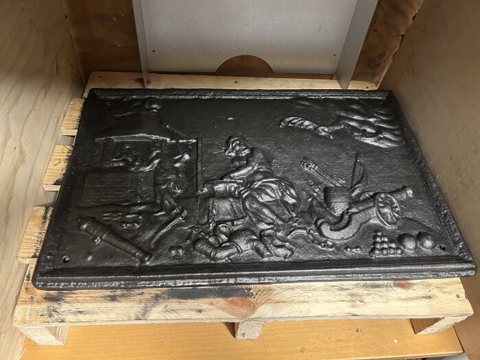 Behandlad braskaminerellief i svart metall på en träpall, detaljer av mänskliga och djurlika figurer avbildade.