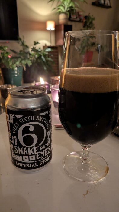 Burk och glas med West Sixth Brewing Snake Eyes Imperial Stout på ett bord, omgivna av växter och tända ljus i bakgrunden.