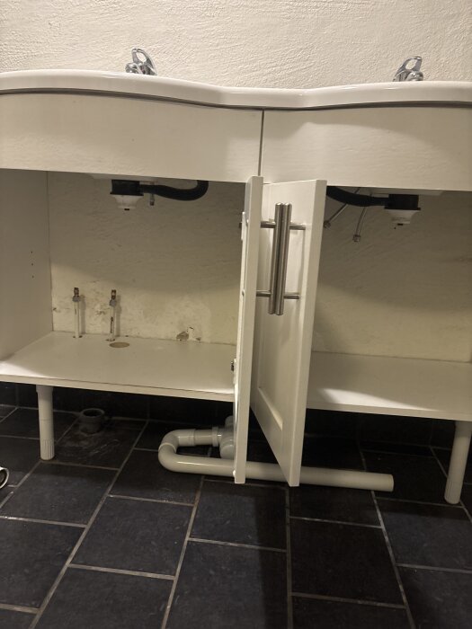Vitt handfatsskåp med öppen dörr, visar rörinstallation under diskbänk, inklusive rör från golv. Mörkt klinkergolv syns.