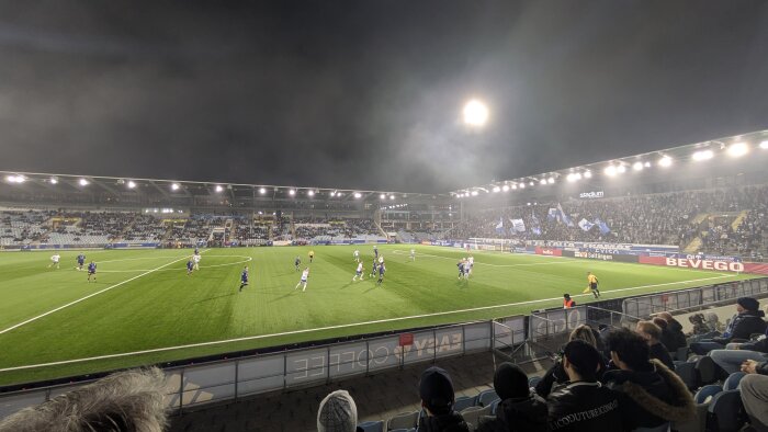 Fotbollsmatch på stadion med publiken på läktaren, spelare i rörelse på planen, och en domare i gul tröja. Kvällstid med strålkastarbelysning.
