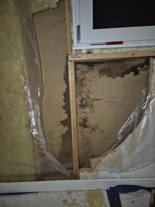 Fukt- och mögelskada bakom fönster, med synliga svarta fläckar på väggen och trasig isolering samt plast. Vattenskador runt fönsteröppningen syns tydligt.