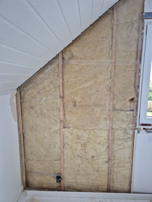 Öppen vägg med synliga reglar och isolering, fuktspår längs kanterna, intill ett delvis synligt fönster.