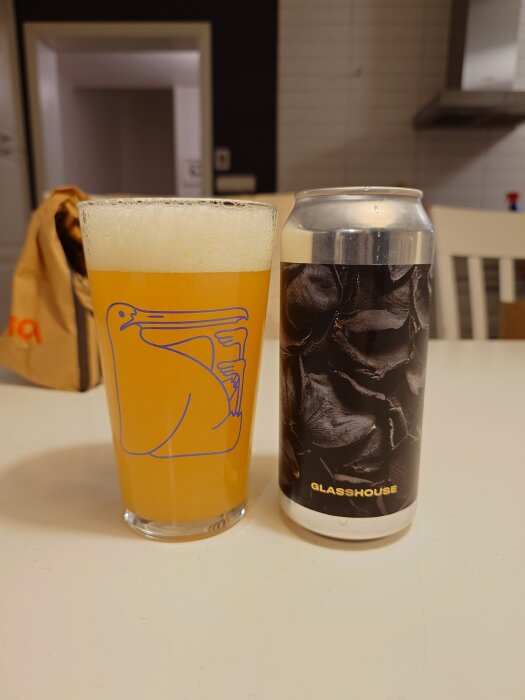Ett glas ljust öl bredvid en burk med etiketten "Glasshouse" på ett vitt bord.