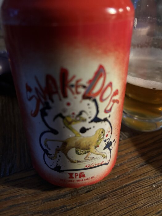 Röd burk med design och texten "Snake Dog" samt "IPA". Bakgrund visar ett glas med öl på ett träbord.