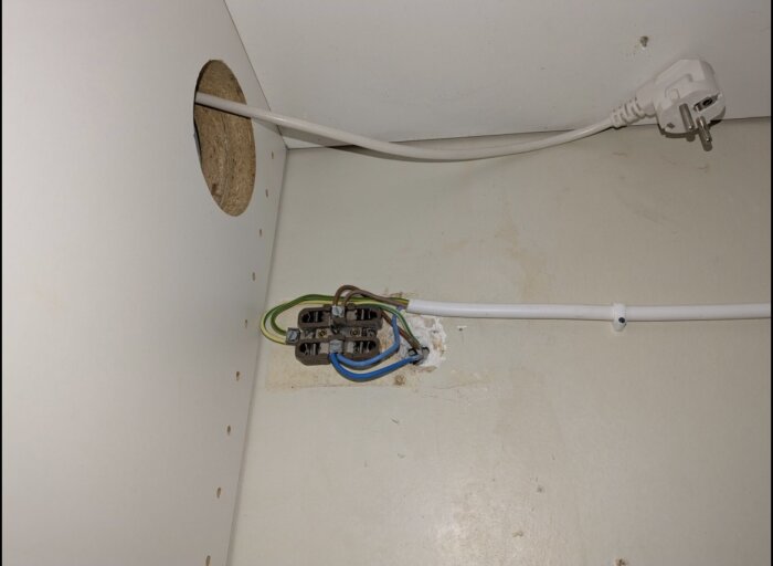 Elinstallation med kabeldragning genom ett hål i väggen, elektriska ledningar i olika färger och kontakt med stickpropp.