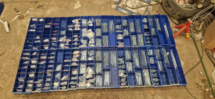 Blå plastbackar fyllda med sorterade skruvar och muttrar i olika storlekar arrangerade på ett golv.