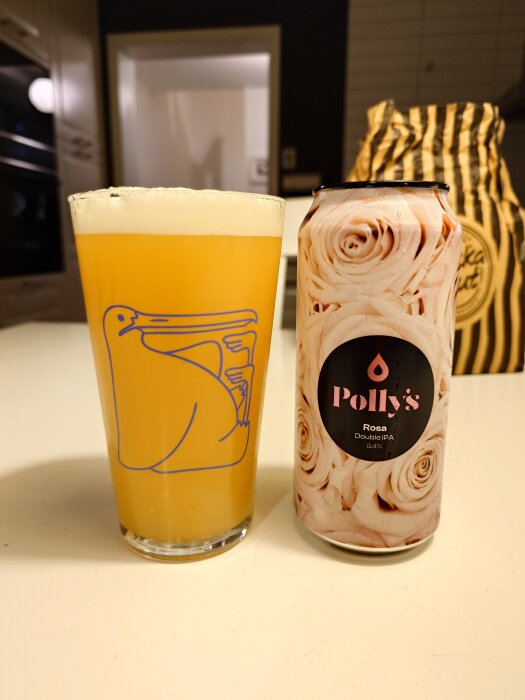 Ölglas och en burk Polly's Rosa Double IPA på ett bord, med en randig kaffeförpackning i bakgrunden.