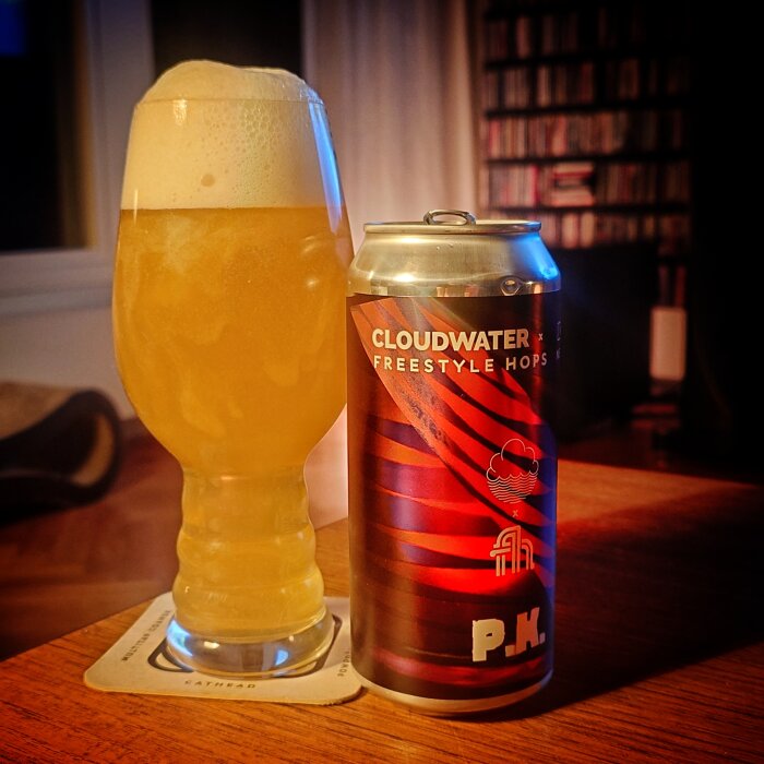 Ett glas ljust öl med skumkrämighet står bredvid en burk från Cloudwater, på ett bord inomhus.