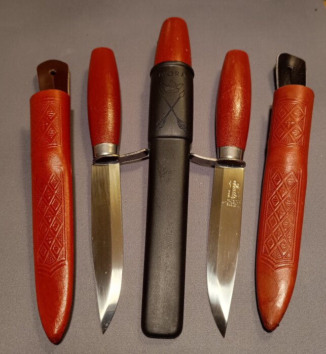 Moraknivar i röda och svarta slidor uppställda i rad, visar traditionell svensk knivdesign med trähandtag och blad av rostfritt stål.
