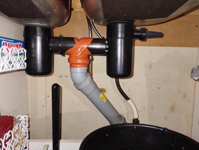Rörsystem under en diskbänk med två diskhoar, inklusive svarta vattenlås och orange kopplingar, del av en villa från 1865 med äldre avloppssystem.