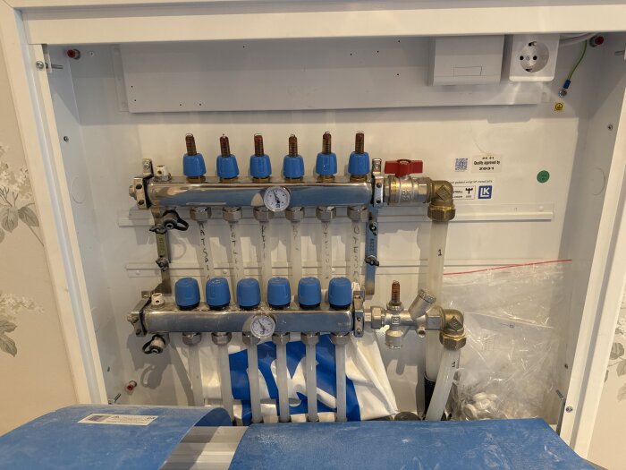 Manifoldskåp för vattenburen golvvärme med blå och vita ventiler, tryckmätare och kopplingar, inuti ett vitt väggskåp.