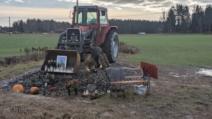 Röd traktor vid ett minnesmärke med blommor, ljus och en bild, omgiven av öppet landskap och en skylt med texten "AXEL".
