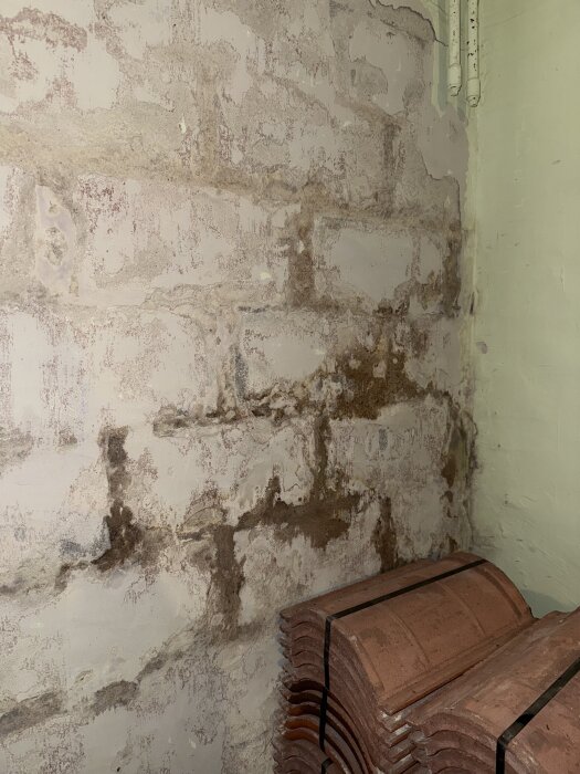 Fuktfläckad källarvägg med avflagad färg och bruna märken, ovanpå takpannor staplade på golvet.