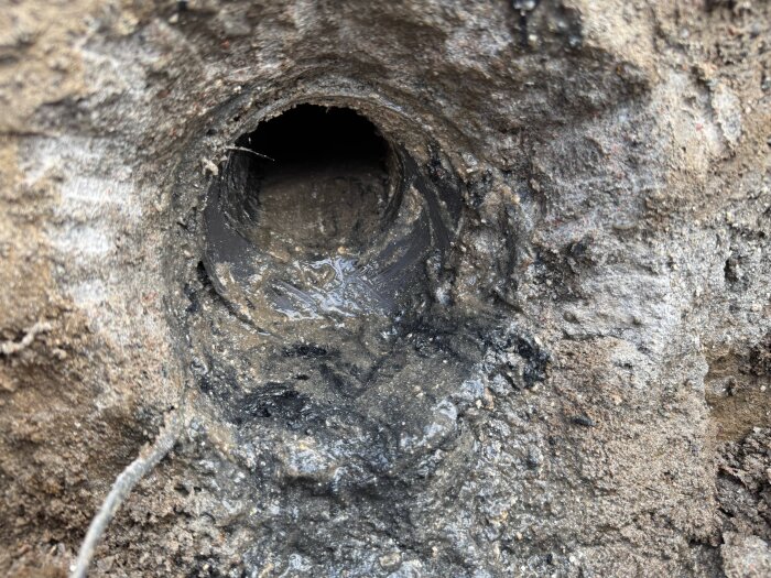 Slamfyllt rör i marken, omgiven av jord och grus, del av avloppssystemet med uppenbara läckage och blockeringar.