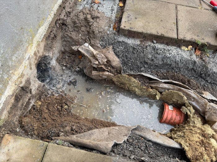 Grävd grop vid husvägg visar exponerat avloppsrör och isolering, omgivet av grus och sand, med synlig vattenansamling.