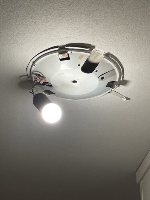 Nedåthängande taklampa med synliga elkablar och en glödande ljuskälla mot ett vitt innertak.