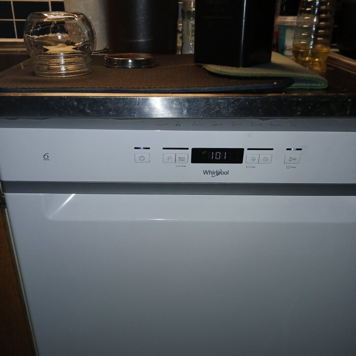 Vit diskmaskin med digital display och kontrollpanel, glasbruk och hushållsartiklar ovanpå.