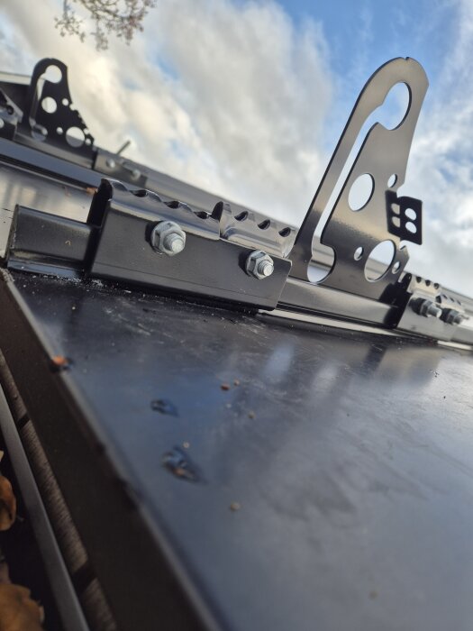 Svart snörasskydd monterat på klicktak, med skruvar fästa nära plåtens kant. Bakgrund av blå himmel och moln syns.