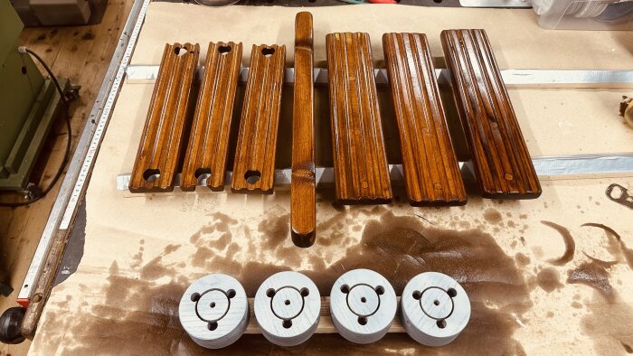 Behandlade trädetaljer och fyra metallfästen på en arbetsbänk under bearbetning för en trampbåt och grannens båt.