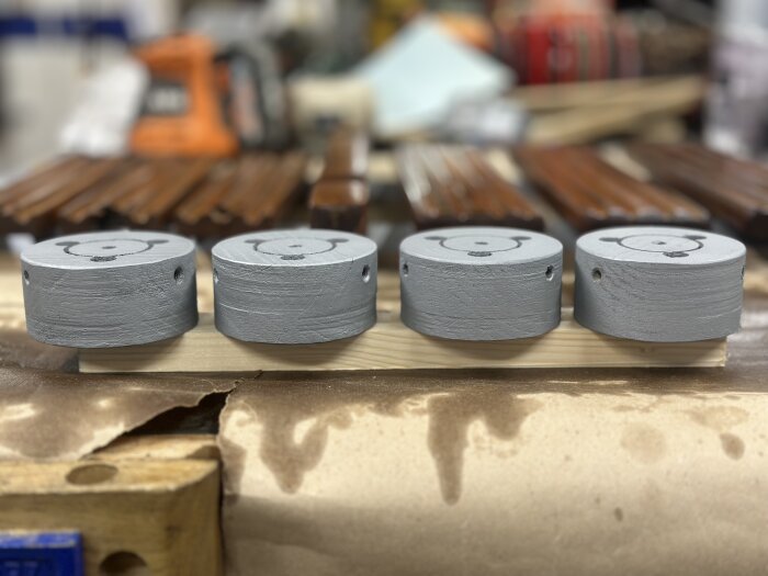 Fyra gråmålade fästen för en trampbåt ligger på en bräda i ett verktygsrum, omgivna av trädetaljer i bakgrunden.