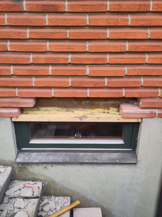 Röd tegelvägg med skador ovanför ett fönster, delar av väggen har spruckit. Byggmaterial och skräp syns vid fönstret och trappan nedanför.