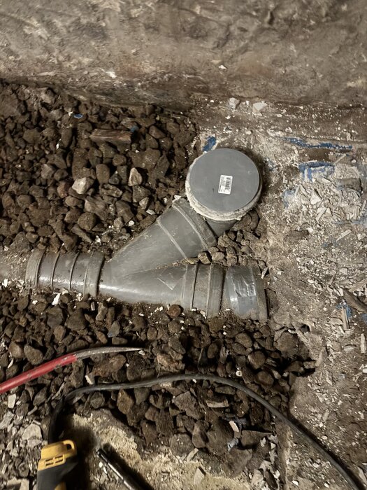 Röranslutning i ett golv med grus och bråte runtomkring, antyder gammal installation med behov av reparation.