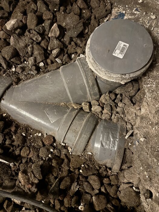 Avloppsrör i golv, delvis täckt av silvertejp och omgiven av grus och jord, tydligt reparationsbehov.