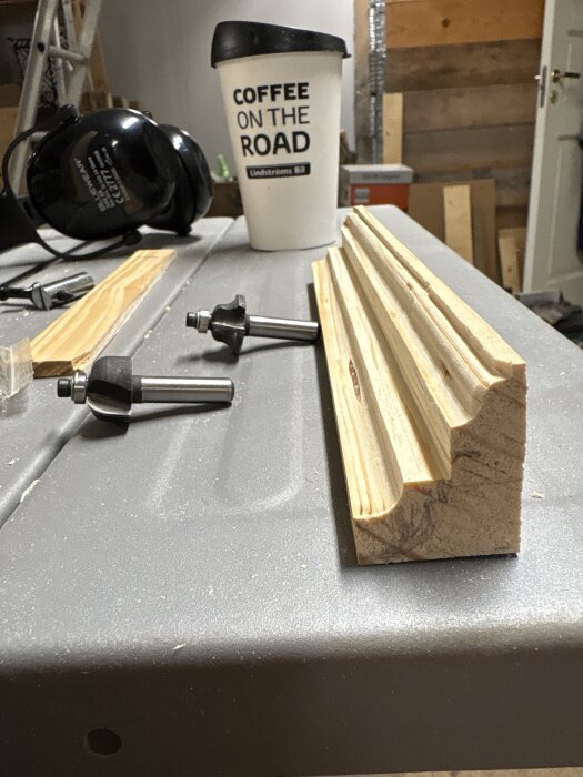 Ett fräst trästycke med dekorativa spår på ett arbetsbord, omgiven av frässtål och en kaffekopp.