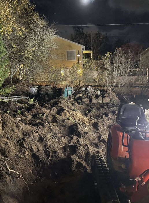 Grävmaskin i arbete på en mörk tomt, omgiven av jordhögar och stenar, med en månbelyst gul byggnad i bakgrunden.