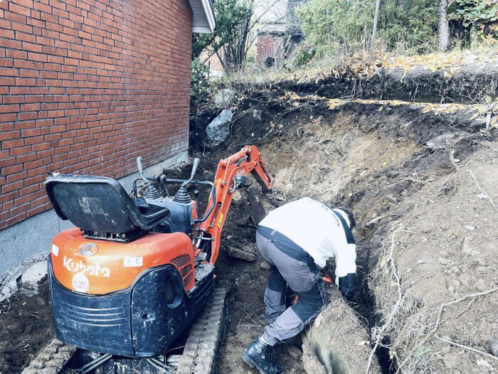 En person gräver med en grävmaskin bredvid en tegelvägg, omgiven av gräs och jord. Grävning nära ett hus för byggprojekt.
