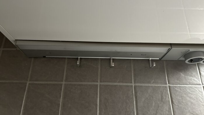 Nedre del av ett köksskåp med utdaterad IKEA Utrusta LED-belysning monterad under, på grå kakelplattor.