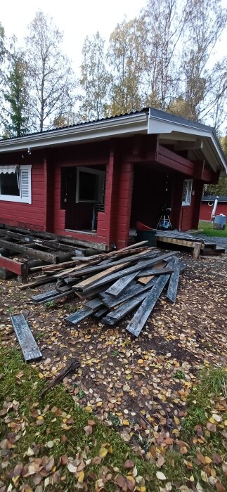 Röd sommarstuga med nedriven altan och brädor på marken i höstlandskap.