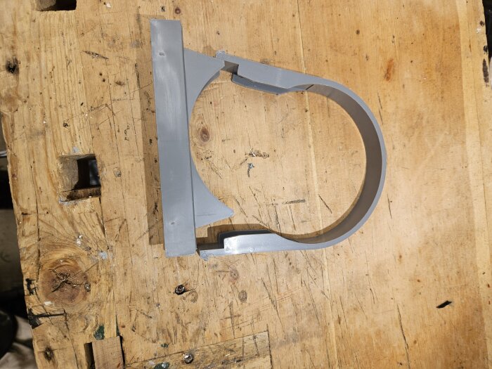 Grå rörklammer av plast ligger på en träyta, används för att montera avloppsrör, möjliggör inställning av distans.